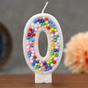 A white candle shaped like the number zero decorated with colorful candy beads for a birthday celebration.
