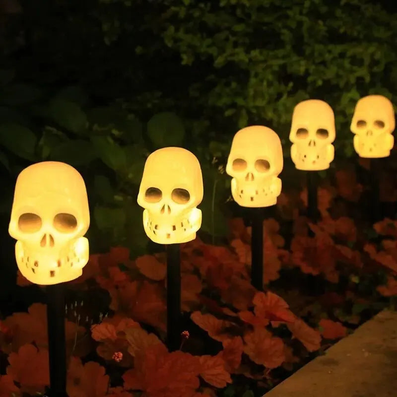 A row of glowing skull solar pathway lights illuminates autumn leaves in a dark garden setting.