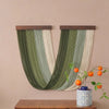 A vertical macrame wall tapestry featuring green and cream stripes hanging on a beige wall.