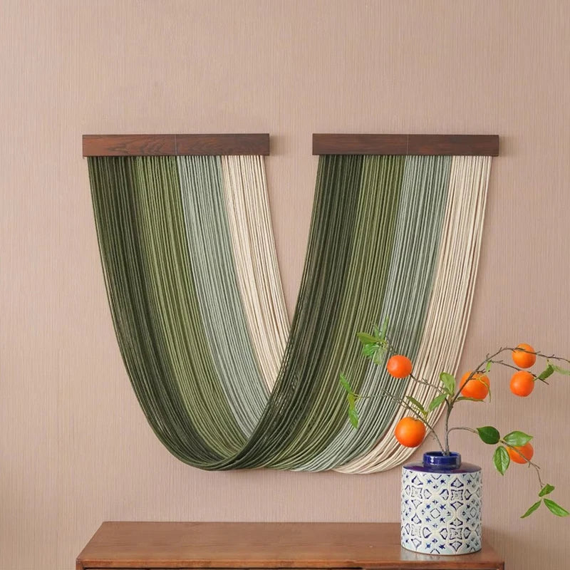 A vertical macrame wall tapestry featuring green and cream stripes hanging on a beige wall.