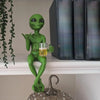 A green alien statue holding a beer mug, displayed on a shelf next to dark books.