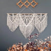 Handwoven boho macrame wall hanging tapestry with white fringe and wooden hanger against a gray wall.