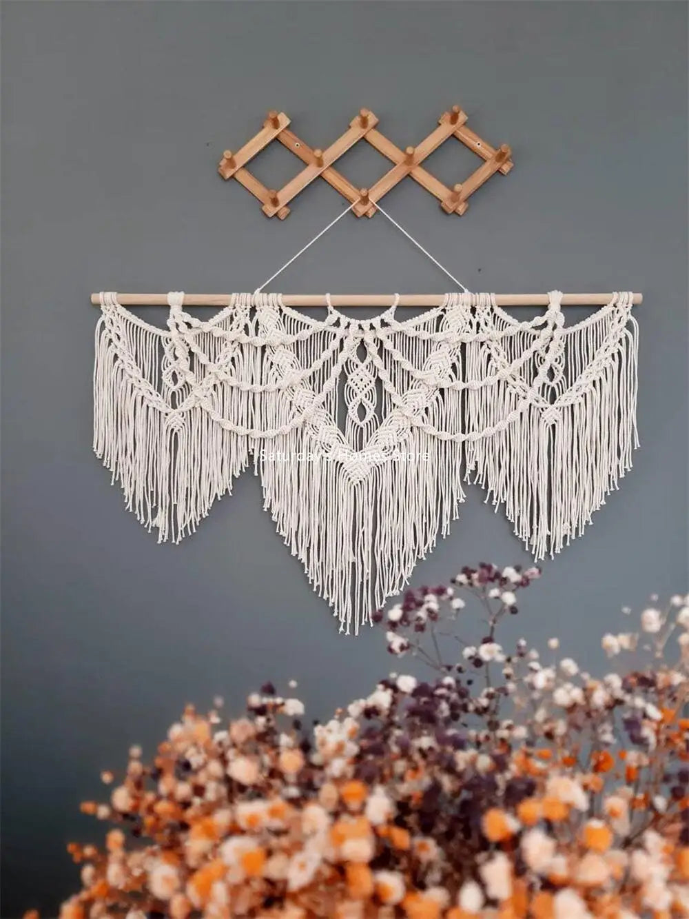 Handwoven boho macrame wall hanging tapestry with white fringe and wooden hanger against a gray wall.