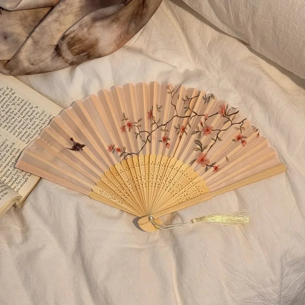 A light wood folding hand fan with delicate floral designs and a black handle resting on white fabric.