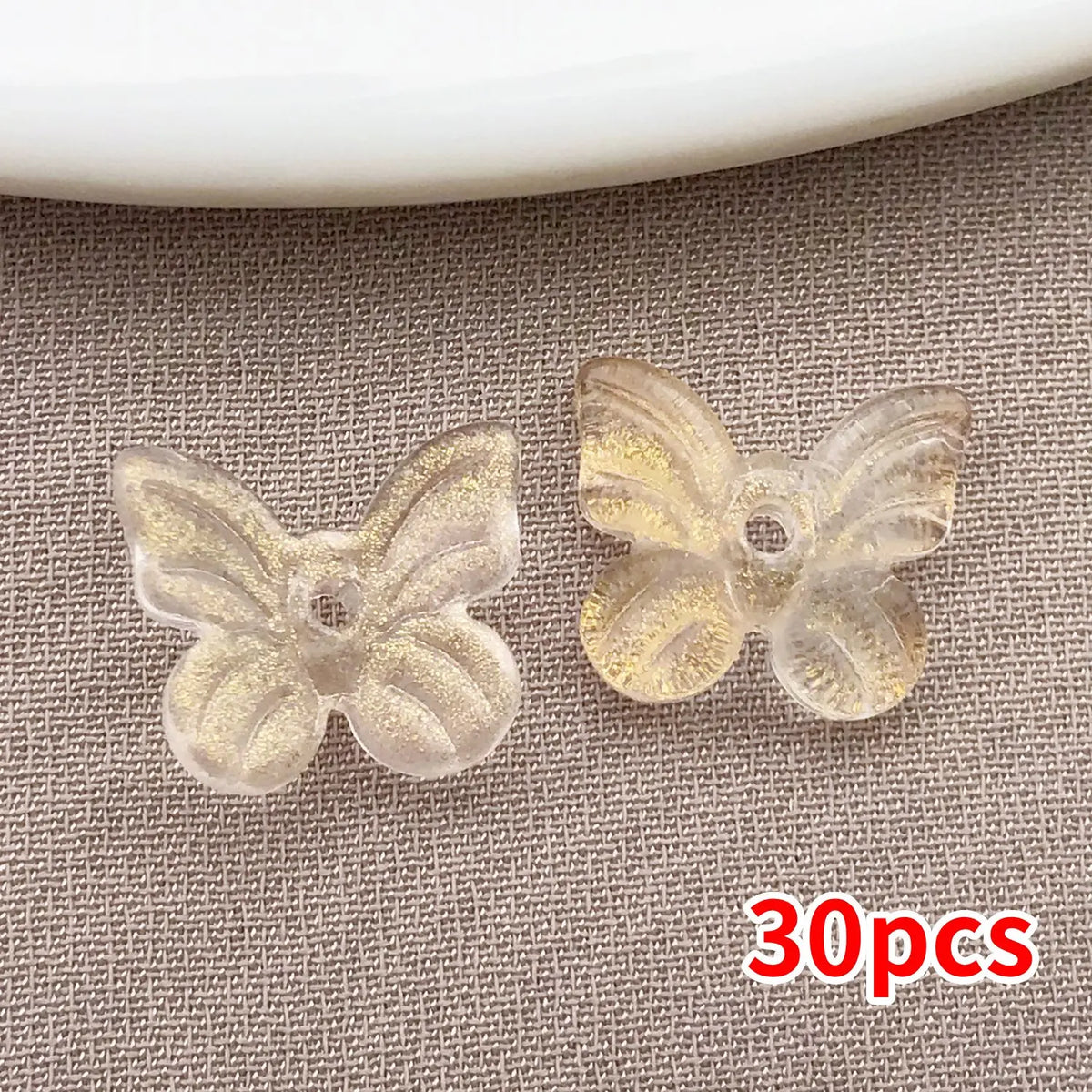 Un juego de 30 cuentas de mariposa de vidrio transparente con acabado de purpurina dorada, mostradas sobre un fondo de tela g