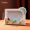 A white photo frame decorated with pink and blue floral blocks and greenery, measuring 7 inches wide.