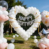 A heart-shaped balloon arch made of white latex balloons with a silver Happy Birthday banner and gold ribbon.