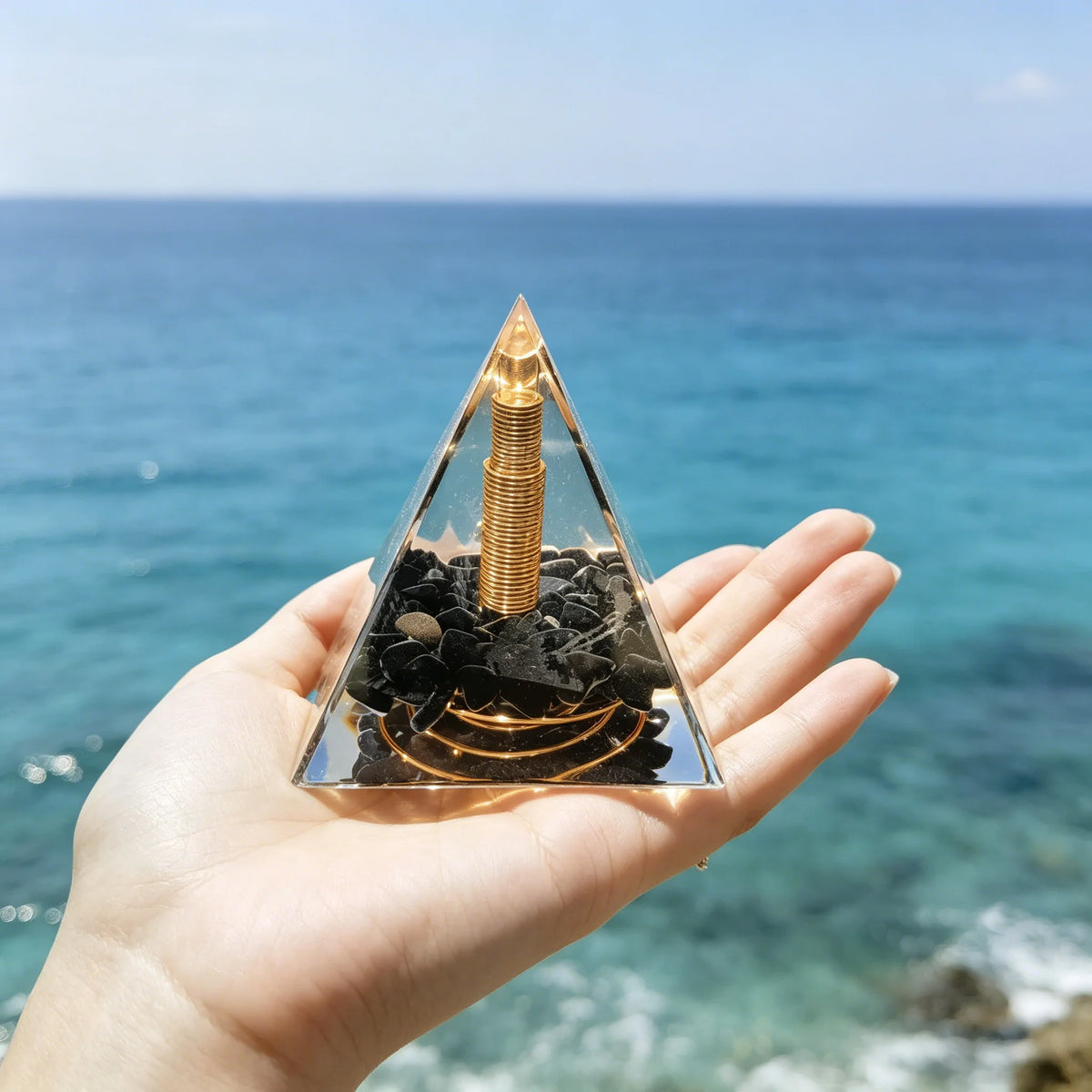 Hand holding a clear resin pyramid with natural black obsidian and a copper coil against a blue ocean background