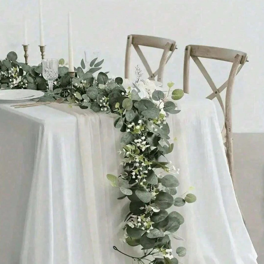 An artificial eucalyptus garland with white flowers draped across a white tablecloth next to wooden chairs.