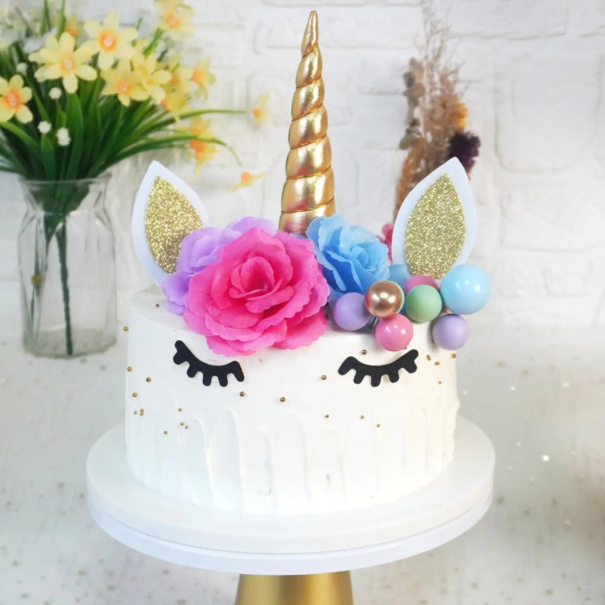 A white unicorn cake decorated with pink roses, blue flowers, gold horn, and glitter accents.