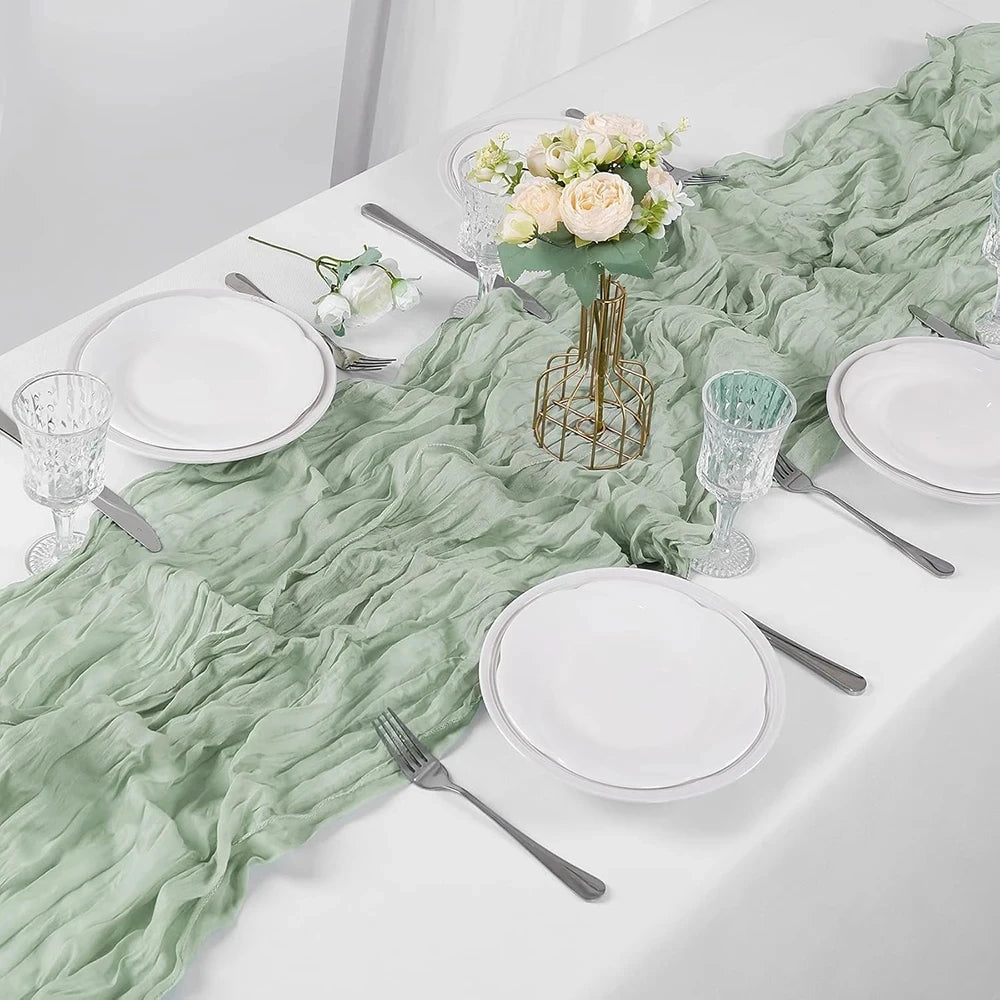 A white table setting featuring a light green sheer gauze table runner, white plates, silverware, and floral centerpiece.