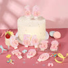 A white layer cake decorated with pink glitter numbers and bows, surrounded by various pink and yellow decorative elements.