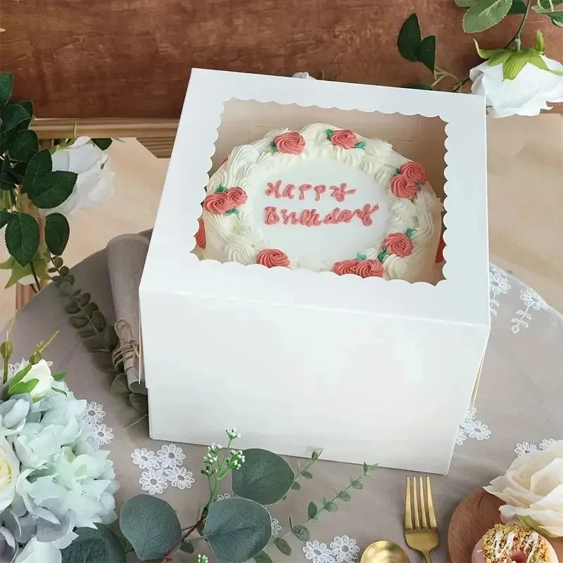 A white bakery box with a window displays a decorated cake featuring pink roses and red lettering.