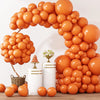 A large archway made of orange latex balloons with white cylindrical stands and loose balloons on a wooden surface.