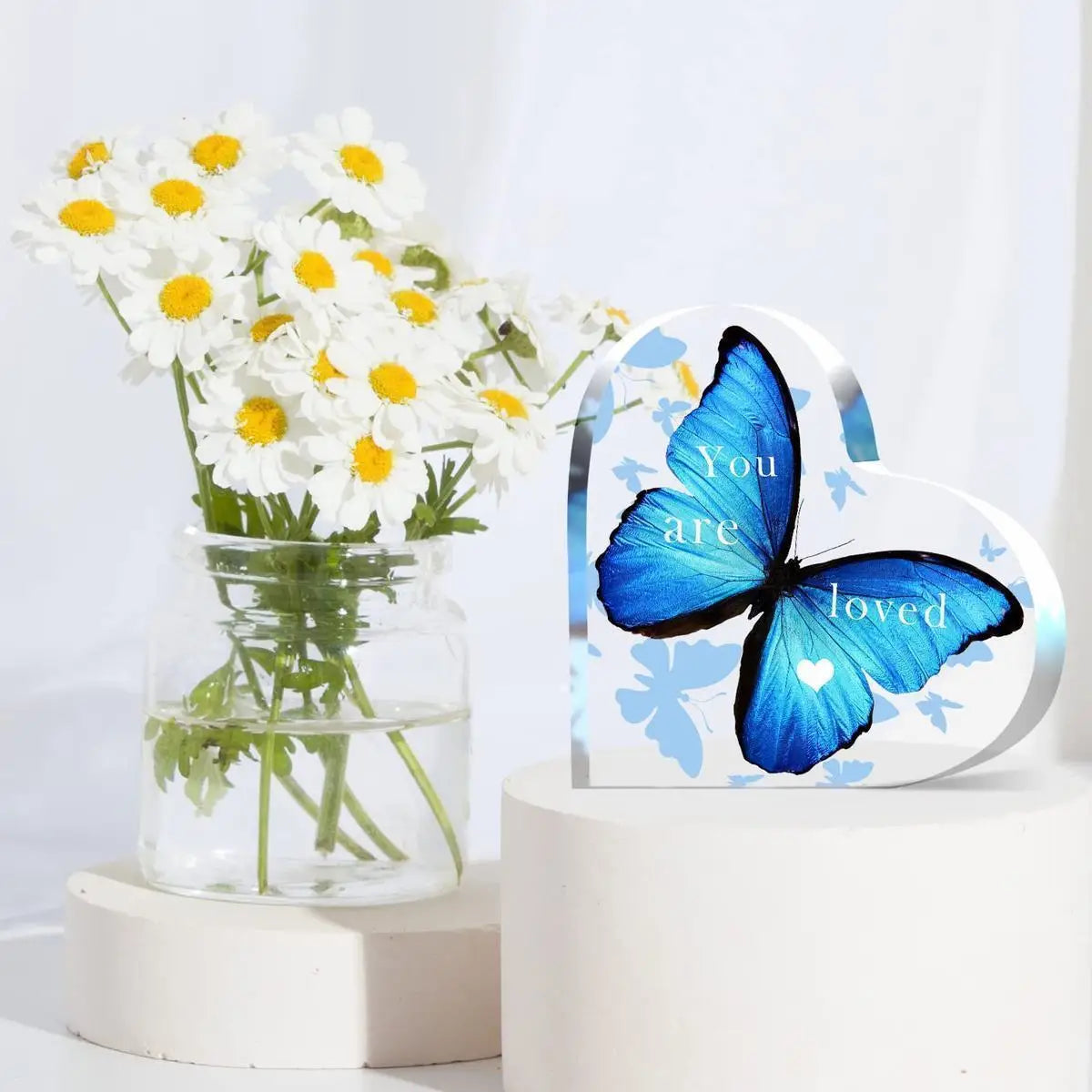 A clear glass vase filled with white daisies sits next to a heart-shaped acrylic sign featuring a blue butterfly and the word