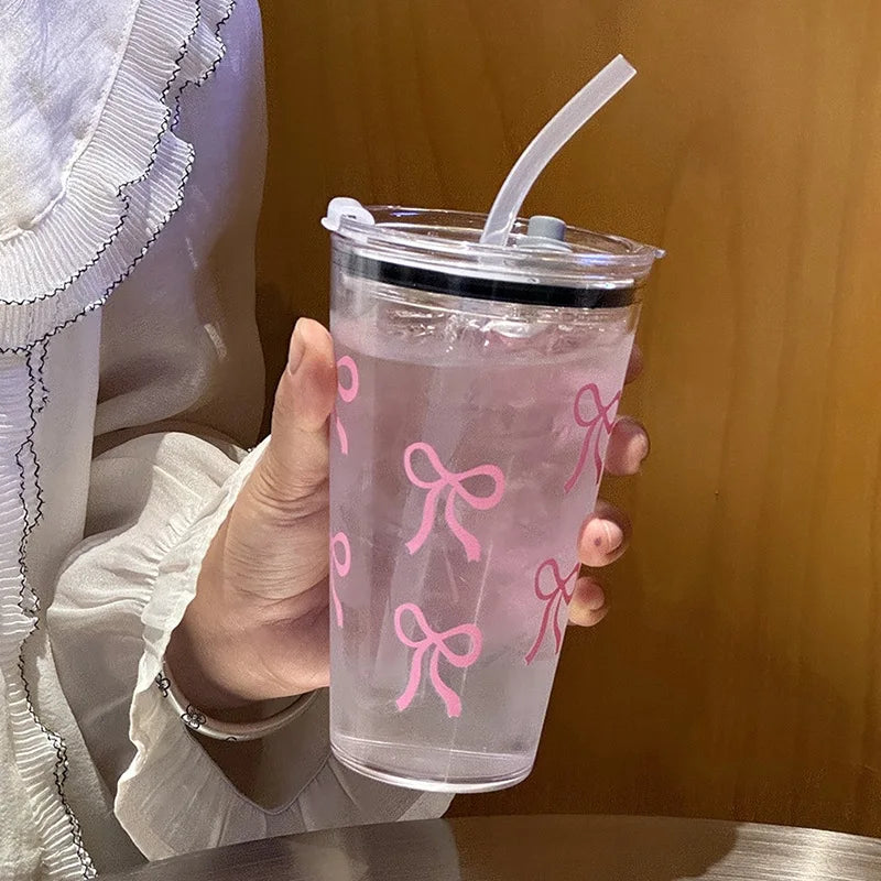 A hand holds a clear pink plastic cup with a lid and straw, decorated with pink bow patterns.