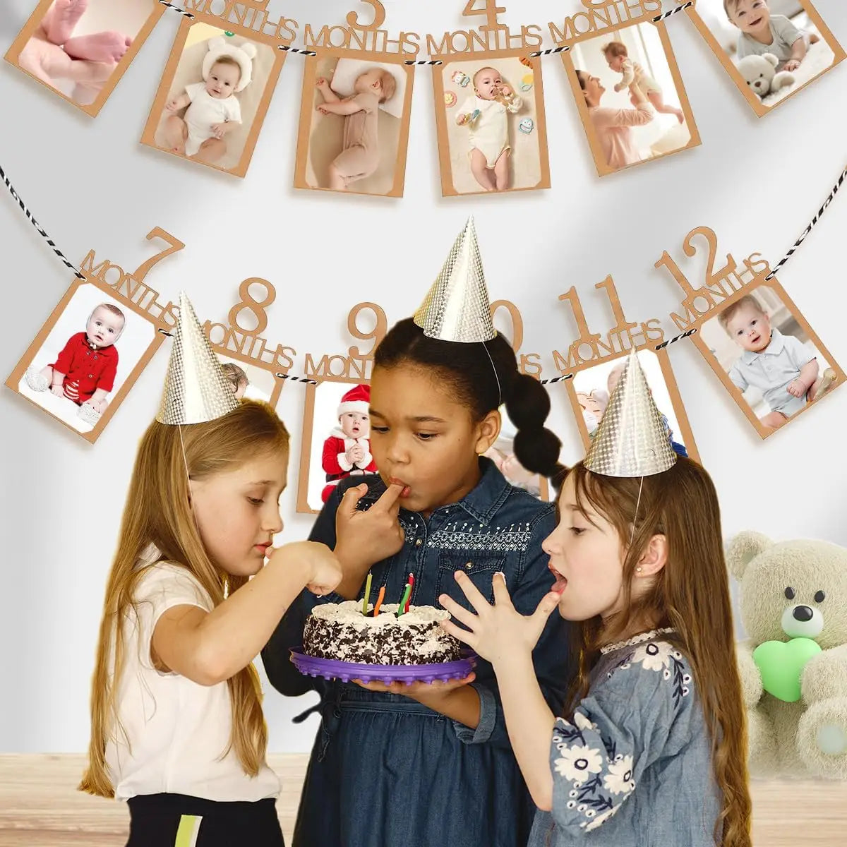 Three young girls interacting with a banner displaying monthly growth records for a baby's first birthday.