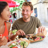 A man and a woman smiling while sharing food served in kraft paper disposable serving boats.