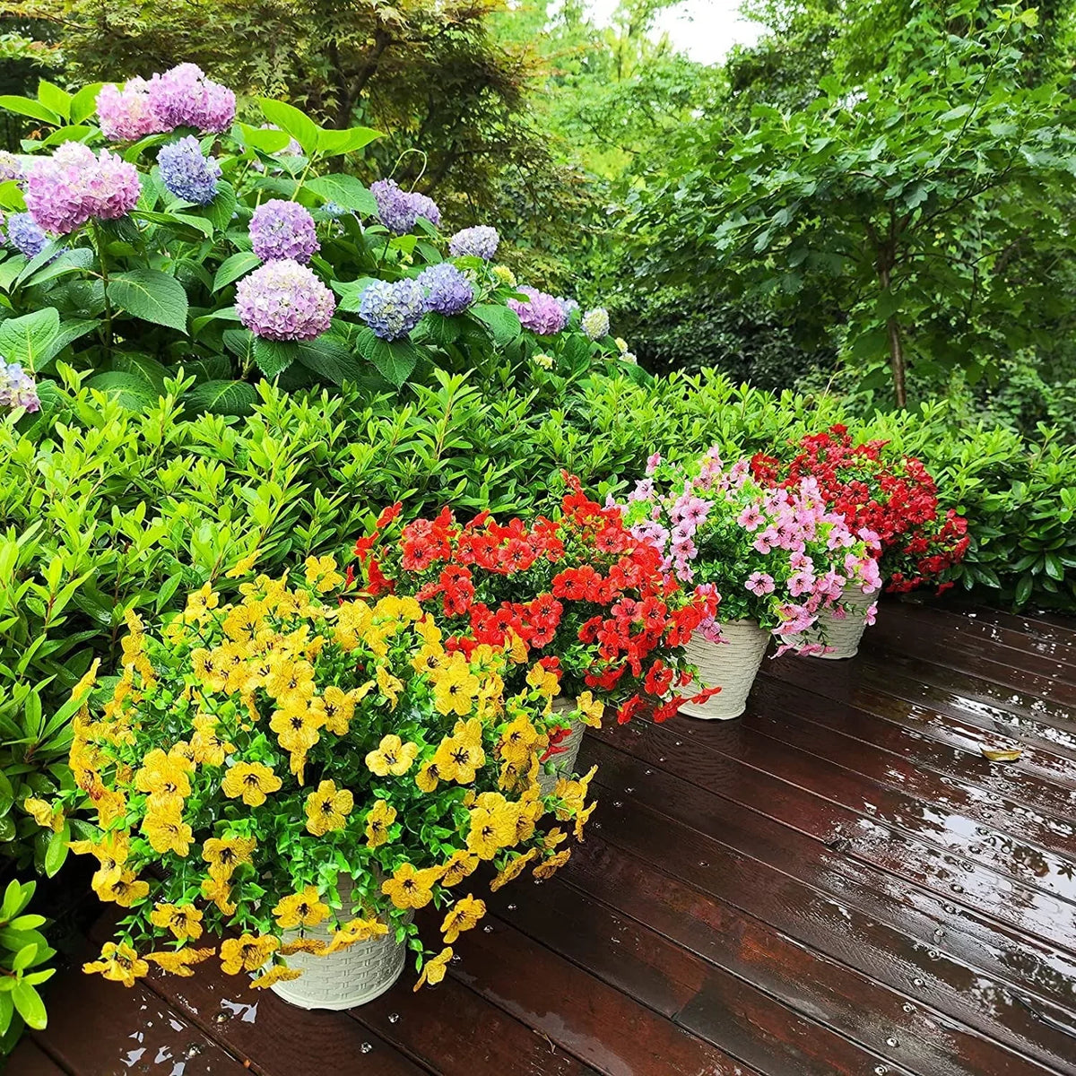 A collection of colorful artificial flowers in various shades including yellow, pink, and red potted on a wooden deck surroun
