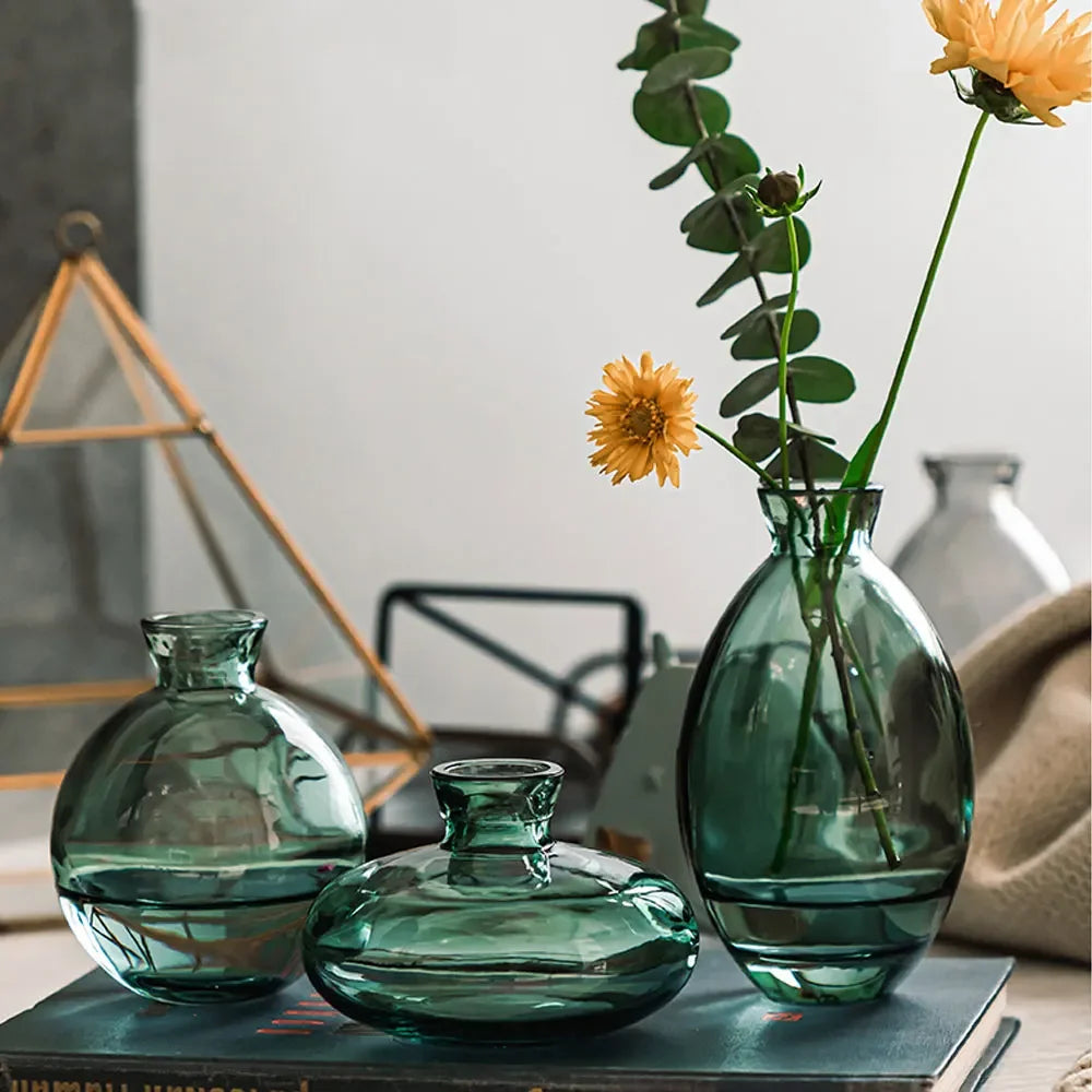A set of three green glass mini vases displayed on a book with a yellow flower in the background.