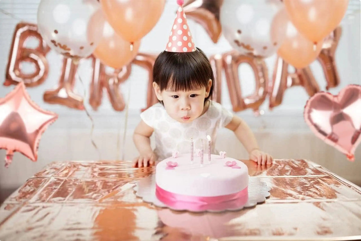 A baby girl is seated in front of a rose gold balloon garland setup for a birthday celebration.