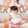 A baby girl is seated in front of a rose gold balloon garland, celebrating a birthday with pink and gold decorations.