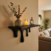 A dark floating shelf holds a vase with greenery and a glowing candle, alongside a small wooden sculpture on a light-colored 
