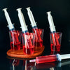 Several clear glass cups hold red liquid and five white party syringes with lids are arranged on a wooden board.