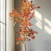 An artificial maple tree with red and orange silk leaves stands in a white pot against a neutral wall.