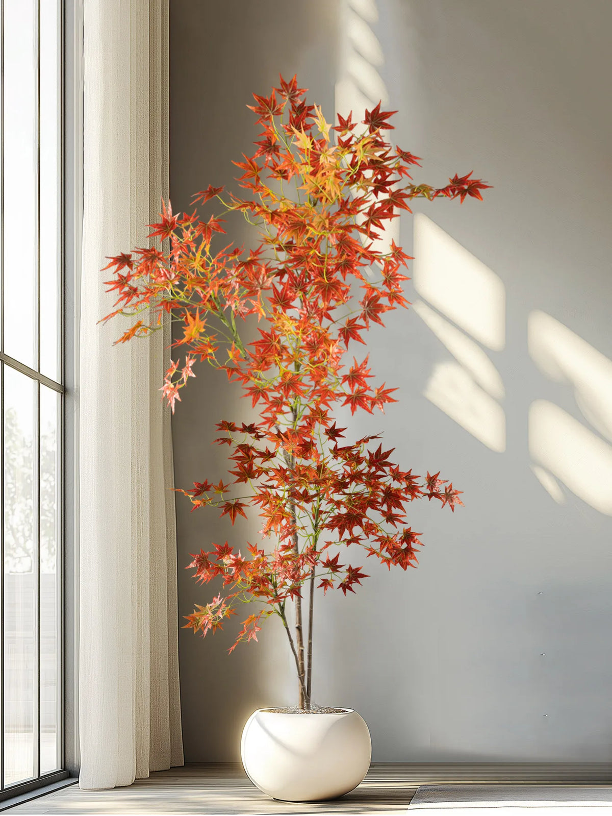An artificial maple tree with red and orange silk leaves stands in a white pot against a neutral wall.