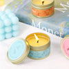 A small jar of yellow candle with a white wick sits on a white surface next to a blue candle and a book.