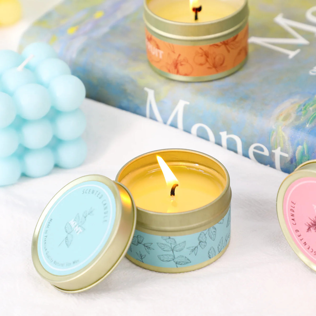 A small jar of yellow candle with a white wick sits on a white surface next to a blue candle and a book.