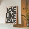 Black text on a white background reads Love Like Jesus framed in wood on a light gray ledge.