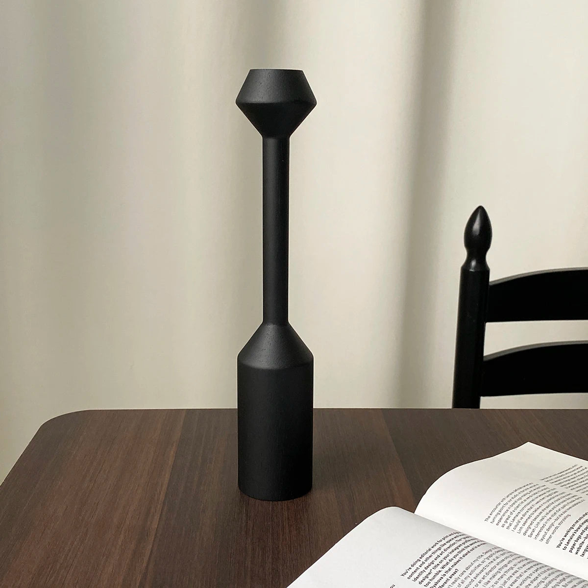 A black, vintage wooden candle holder with a flared top and cylindrical base sits on a dark wooden table.