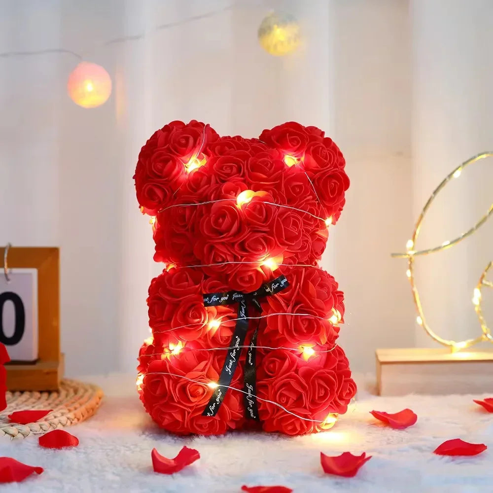A bright red artificial rose teddy bear with glowing lights sits on a white surface surrounded by scattered red rose petals.