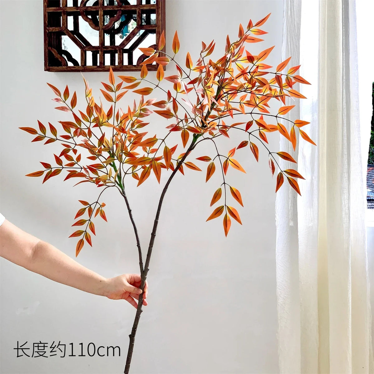 A tall artificial Nandina plant with orange-red leaves is displayed against a white wall next to a window.