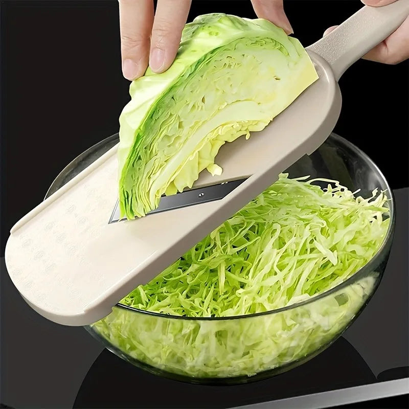A hand is using a manual cabbage shredder with stainless steel blades to shred a head of green cabbage into a glass bowl.