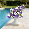A white and purple artificial petunia flower arrangement in a white pedestal vase placed on a stone patio beside a swimming p