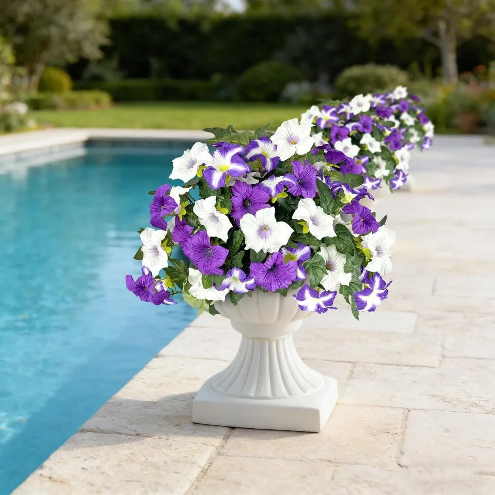 A white and purple artificial petunia flower arrangement in a white pedestal vase placed on a stone patio beside a swimming p