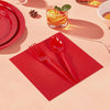 A red paper party tableware set featuring dinner plates, napkins, cups, and cutlery arranged on a light pink surface.