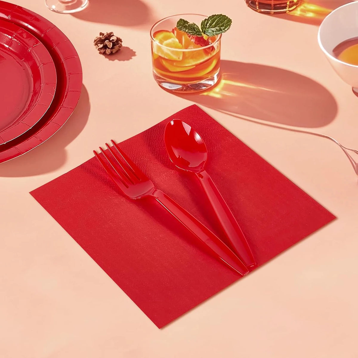 A red paper party tableware set featuring dinner plates, napkins, cups, and cutlery arranged on a light pink surface.