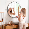 A woman is looking in a large round vanity mirror with a matte metal frame in a bright bathroom setting.