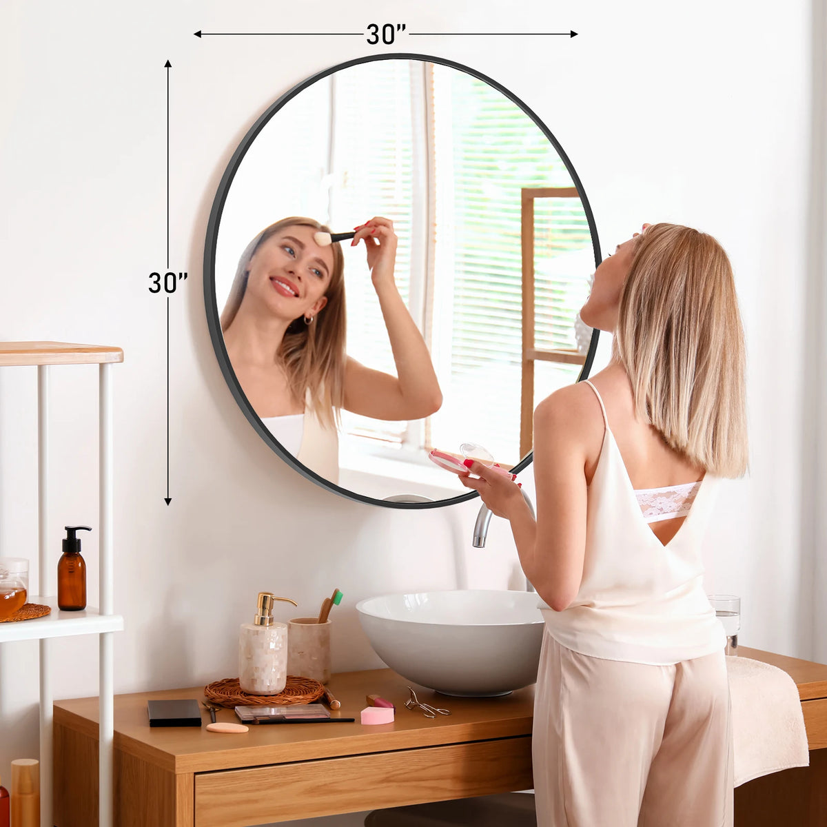 A woman is looking in a large round vanity mirror with a matte metal frame in a bright bathroom setting.