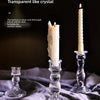 A tall, clear glass pillar candle holder with a white candle is displayed on a gray fabric.