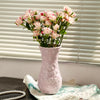 A pink ceramic vase filled with pink roses sits on a dark surface next to a white cloth.