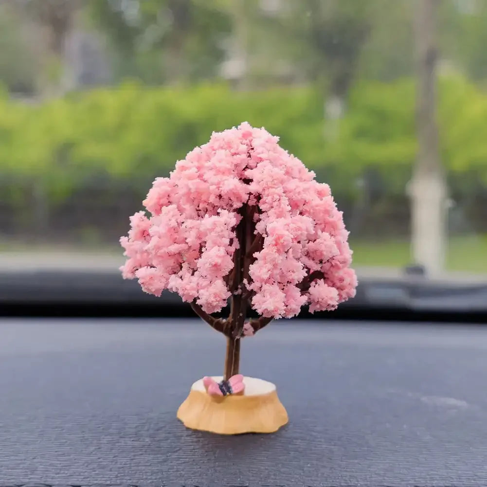 A miniature tree ornament with pink flowers on a wooden base displayed on a dark dashboard.
