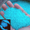 A hand holds a pile of bright cyan glow-in-the-dark luminous pebbles for garden and aquarium decor.