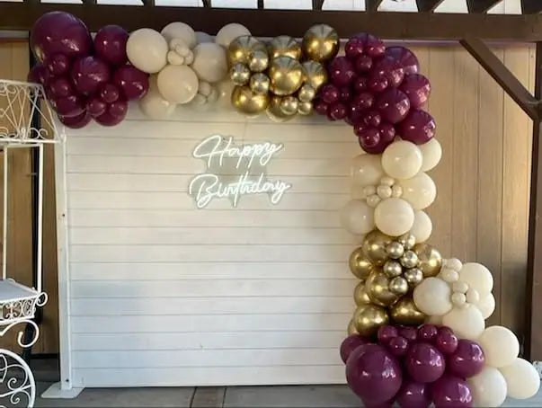 A balloon garland arch featuring burgundy, gold, and white balloons decorating a white wooden backdrop.