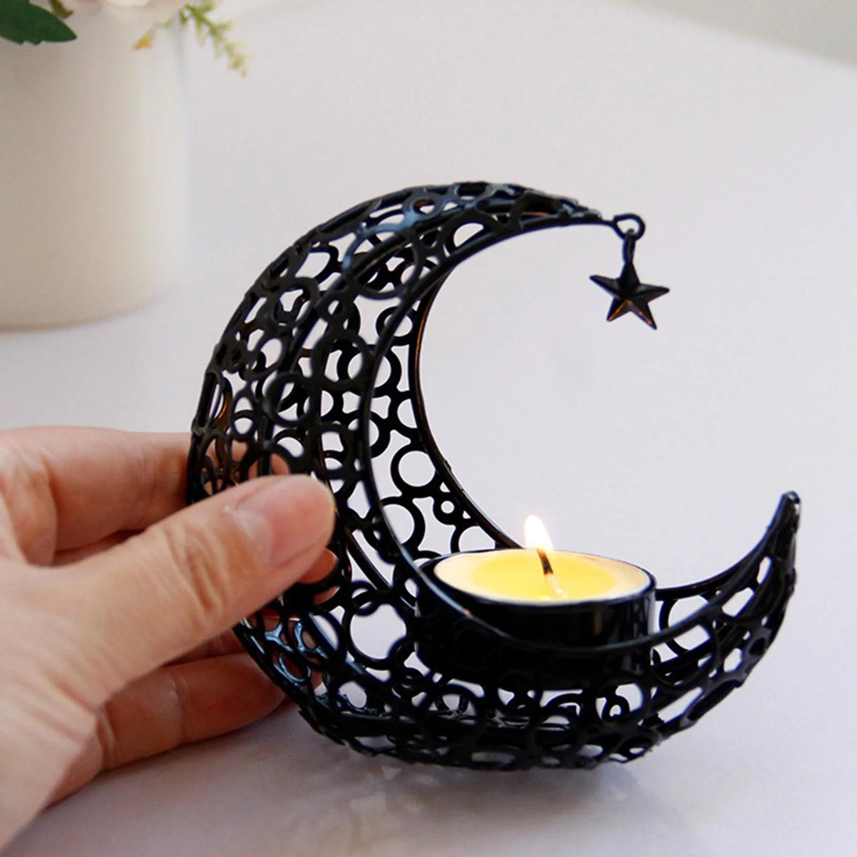 A hand holds a black, intricate crescent moon candle holder with a lit candle and a small star decoration.