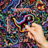 Une main tient un set de stickers de dinosaures et d'animaux lumineux sur un fond abstrait coloré.